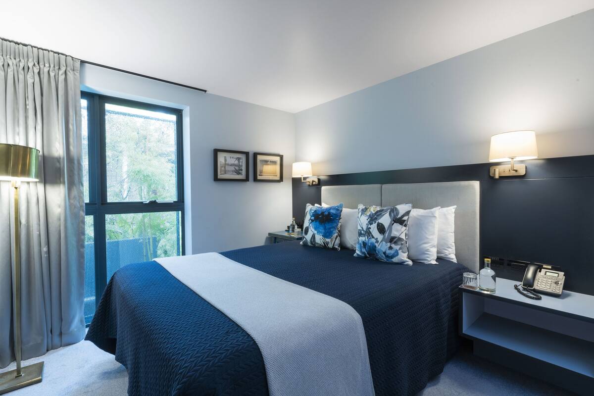 Une chambre d’hôtel moderne avec un grand lit, une table de chevet, une lampe et une fenêtre avec des rideaux.