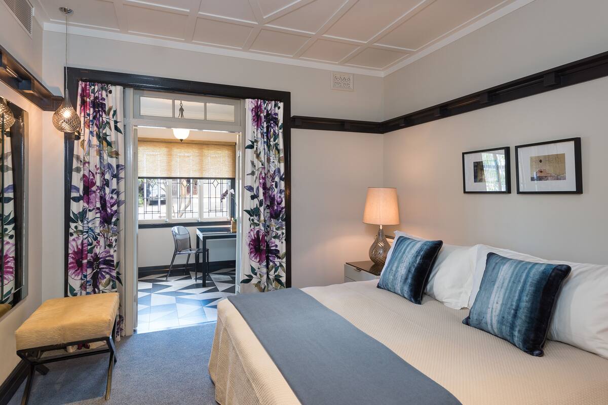 Une chambre d’hôtel avec un lit, un bureau et une chaise. Il y a une fenêtre avec des rideaux, une lampe et deux tableaux encadrés au mur.