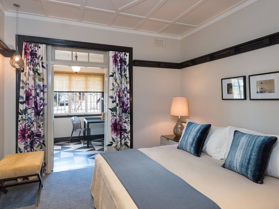 Ein Hotelzimmer mit einem Bett, einem Schreibtisch und einem Stuhl. Es gibt ein Fenster mit Vorhängen, eine Lampe und zwei gerahmte Bilder an der Wand.