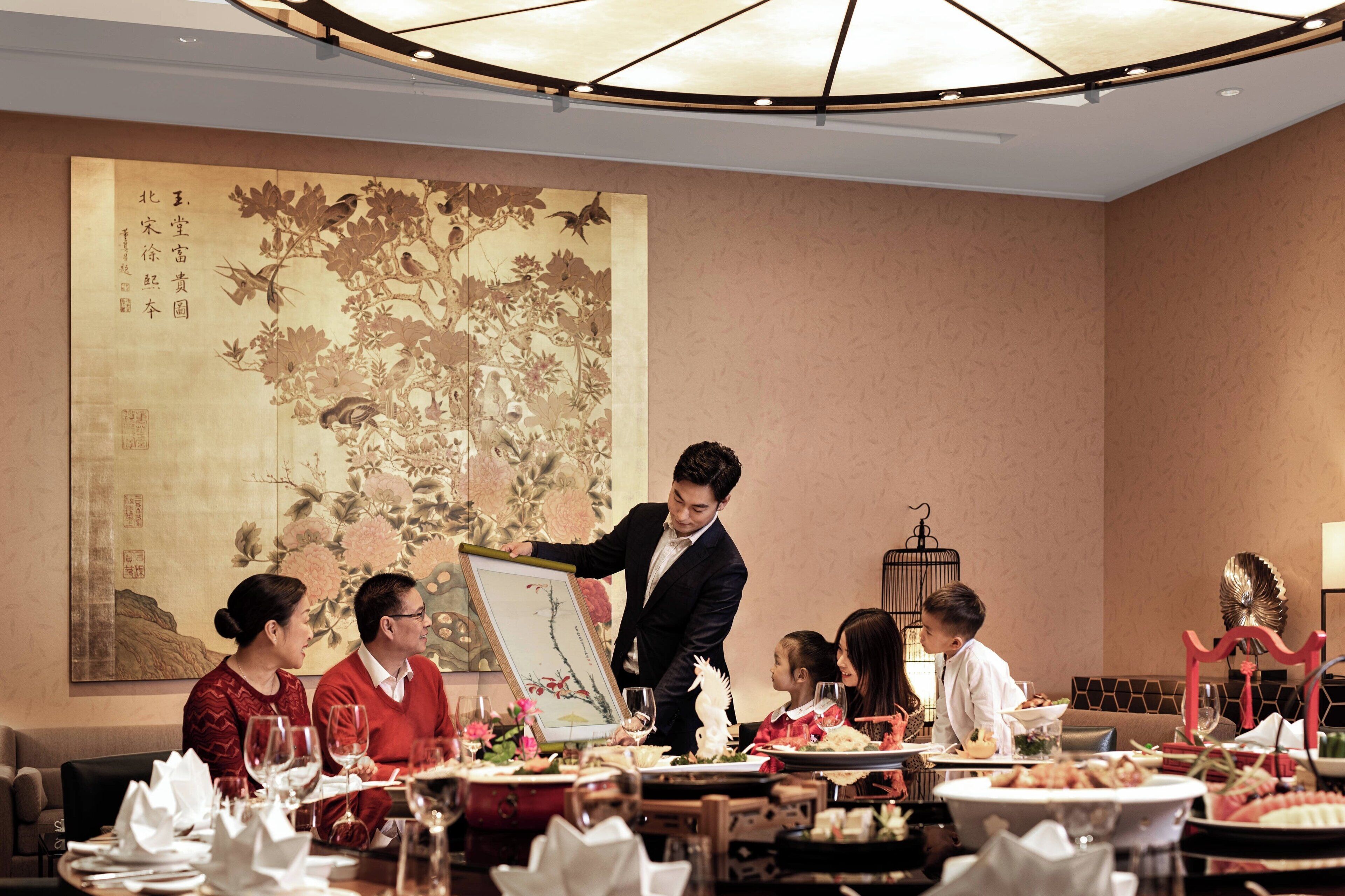 A dining area with a traditional Chinese painting on the wall, a waiter serving food, and guests seated at tables with various dishes and drinks.