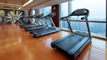 Ein Fitnessraum mit Laufbändern, die an einer Wand mit großen Fenstern mit Blick auf eine Stadtansicht angeordnet sind.