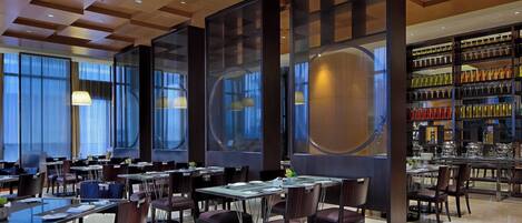 A modern dining area with tables and chairs, large windows, and a decorative wall with shelves.