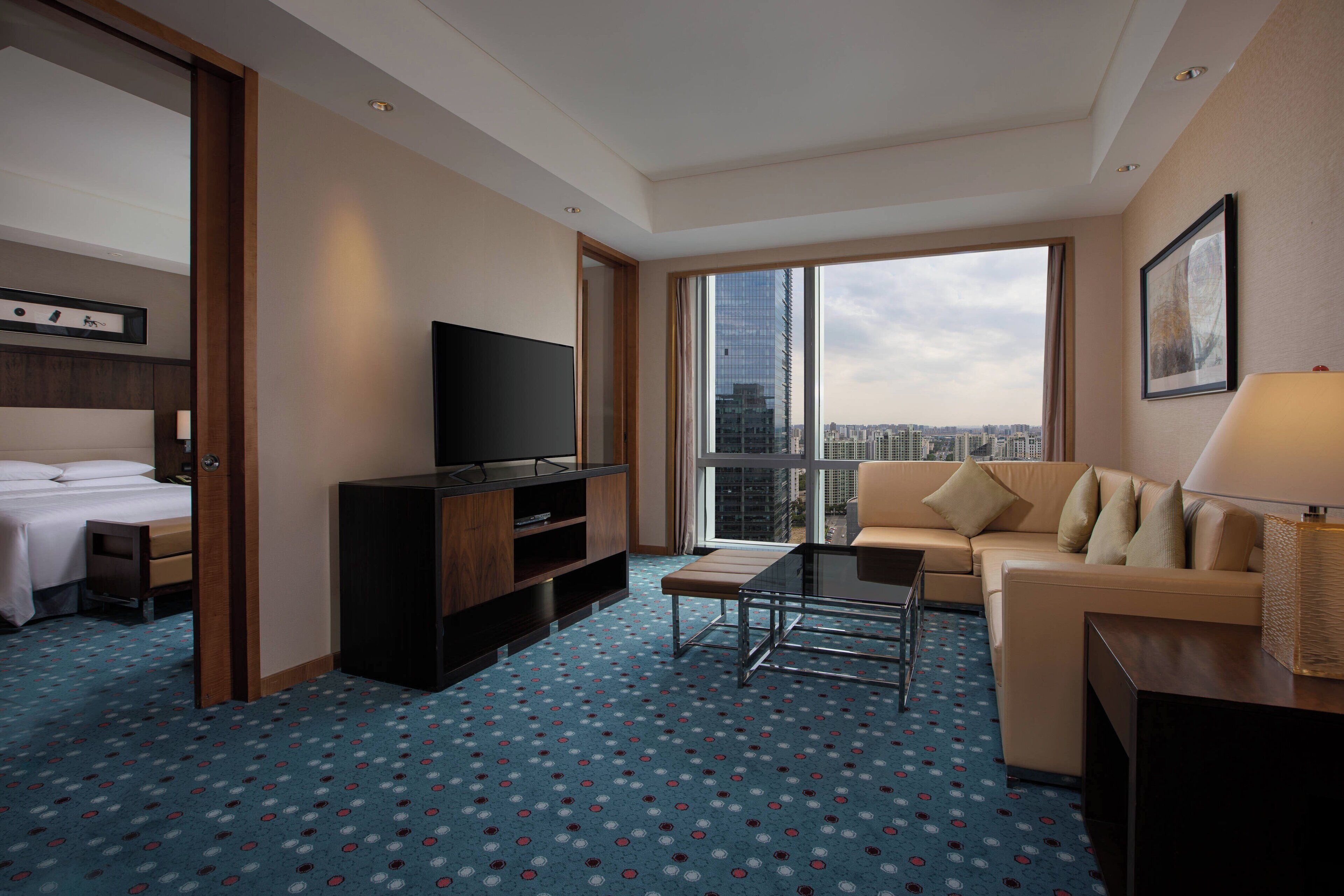 A hotel room with a bed, a TV, a sofa, a coffee table, and a large window with a city view.