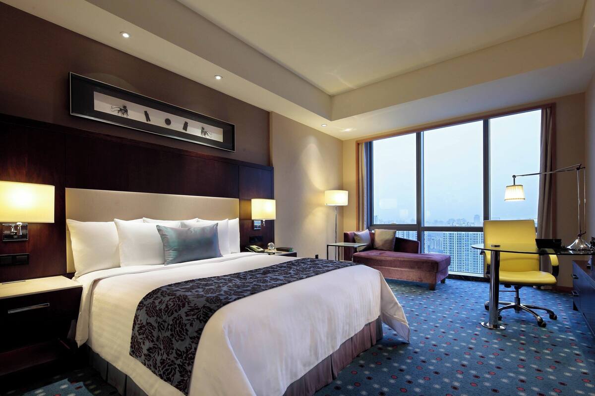 Habitación de hotel con una cama grande, dos mesitas de noche con lámparas, un escritorio con silla, un pequeño sofá y un ventanal con vistas a la ciudad.