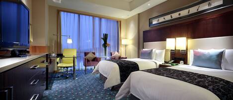 A hotel room with two beds, a desk with a TV, a chair, and a large window with curtains.
