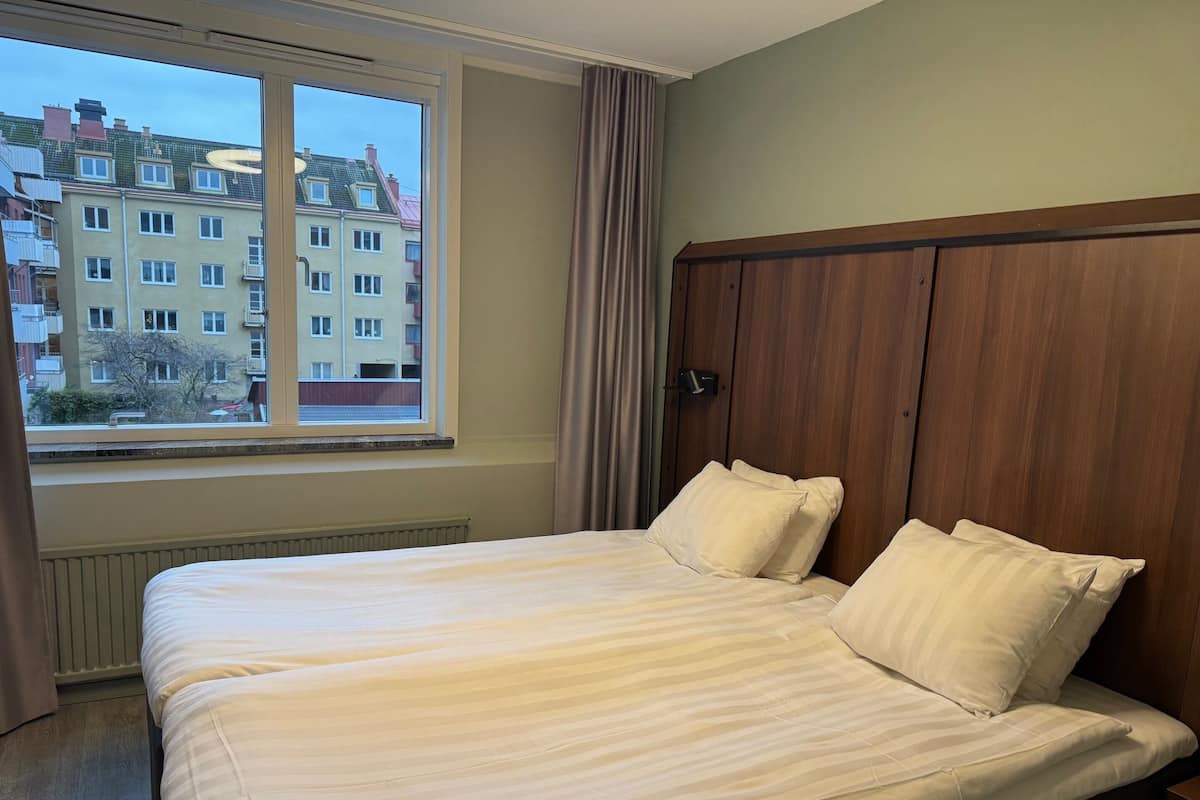 A hotel room with a large bed, a wooden headboard, a window with a view of buildings, and a radiator under the window.