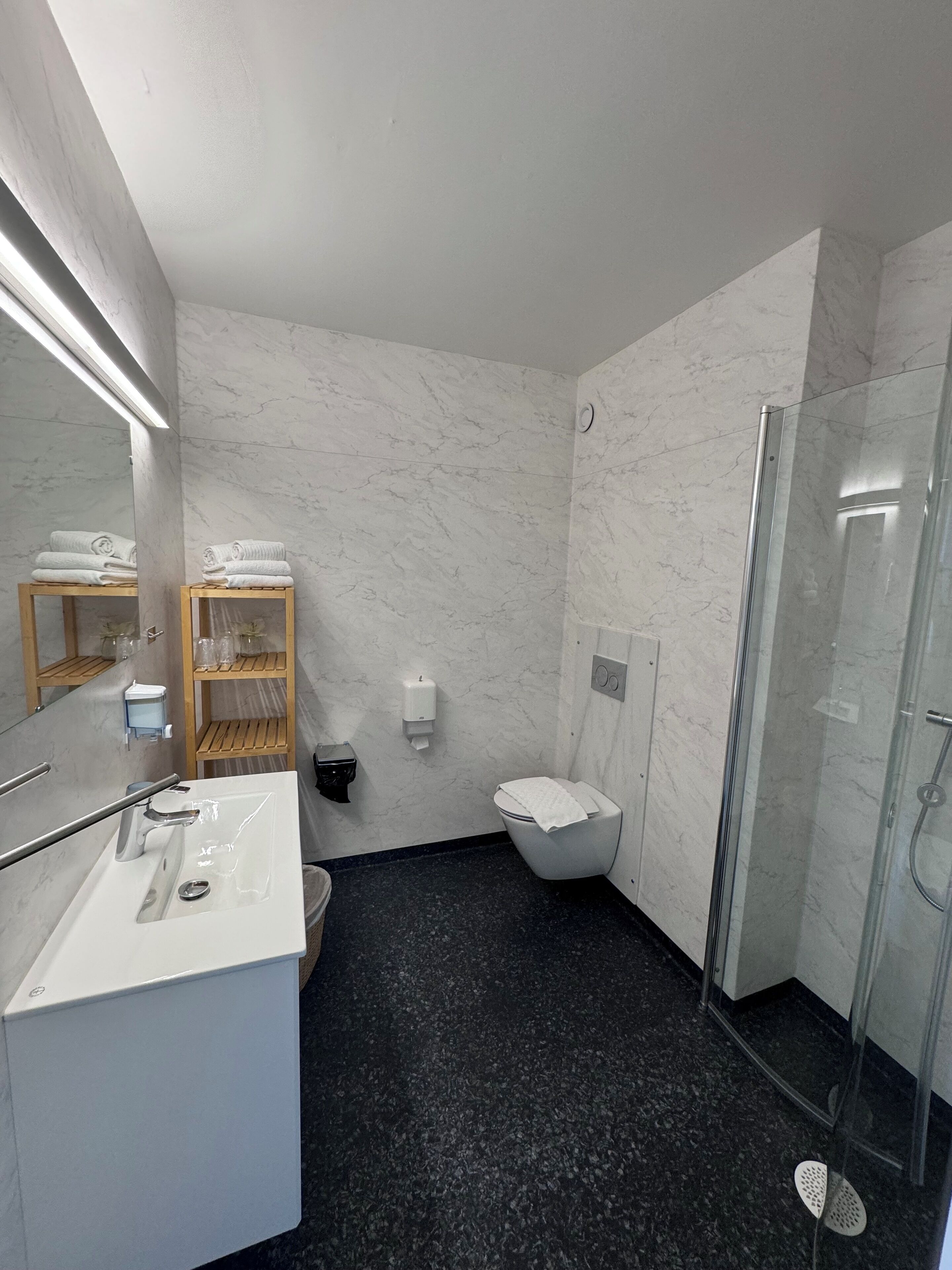 A modern bathroom with a marble wall, a glass shower enclosure, a white sink, and a wooden shelf with towels.