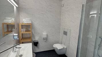 A modern bathroom with a marble wall, a glass shower enclosure, a white sink, and a wooden shelf with towels.