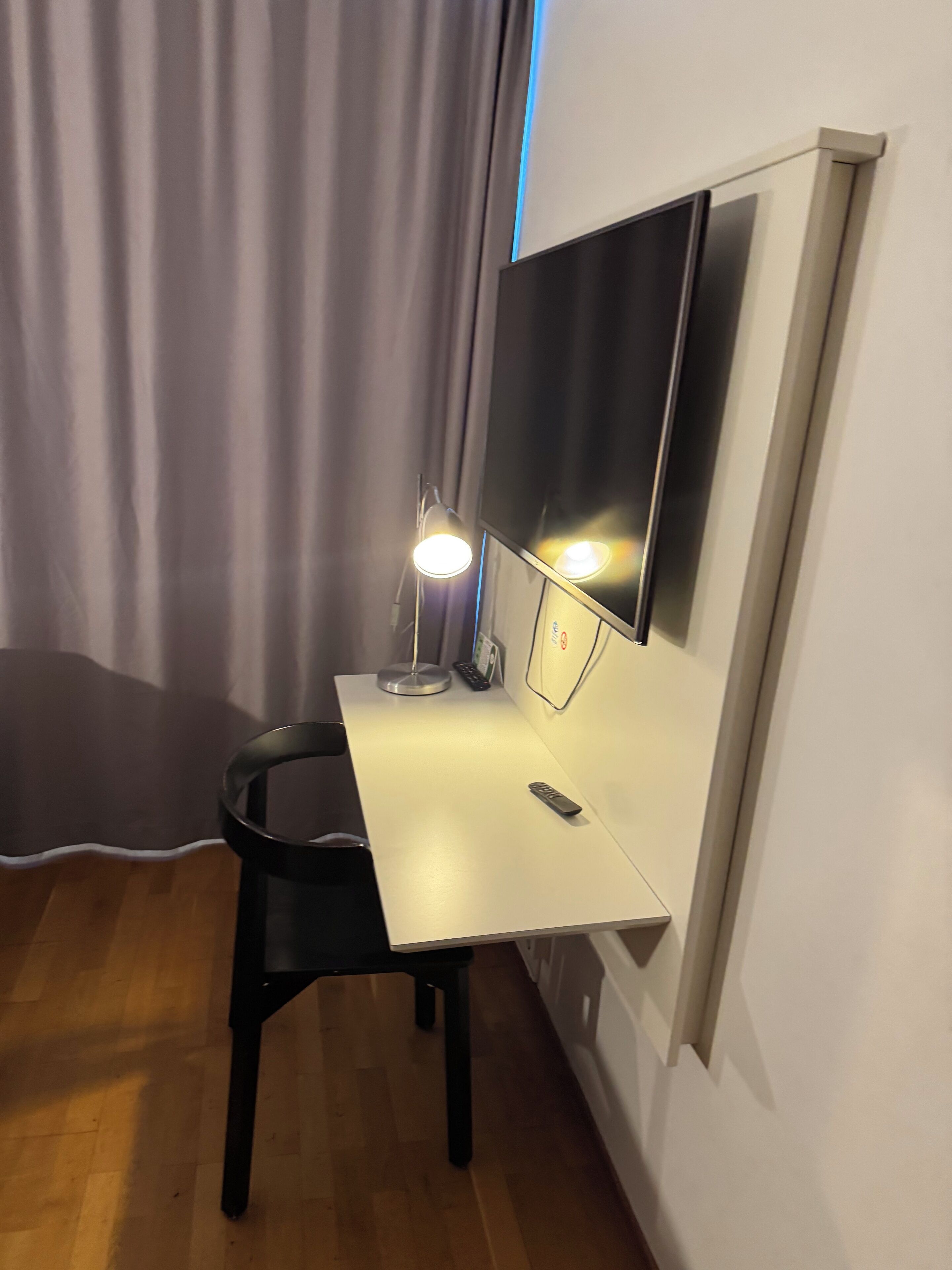 A small, modern room with a white desk, a black chair, a desk lamp, and a flat-screen TV mounted on the wall.