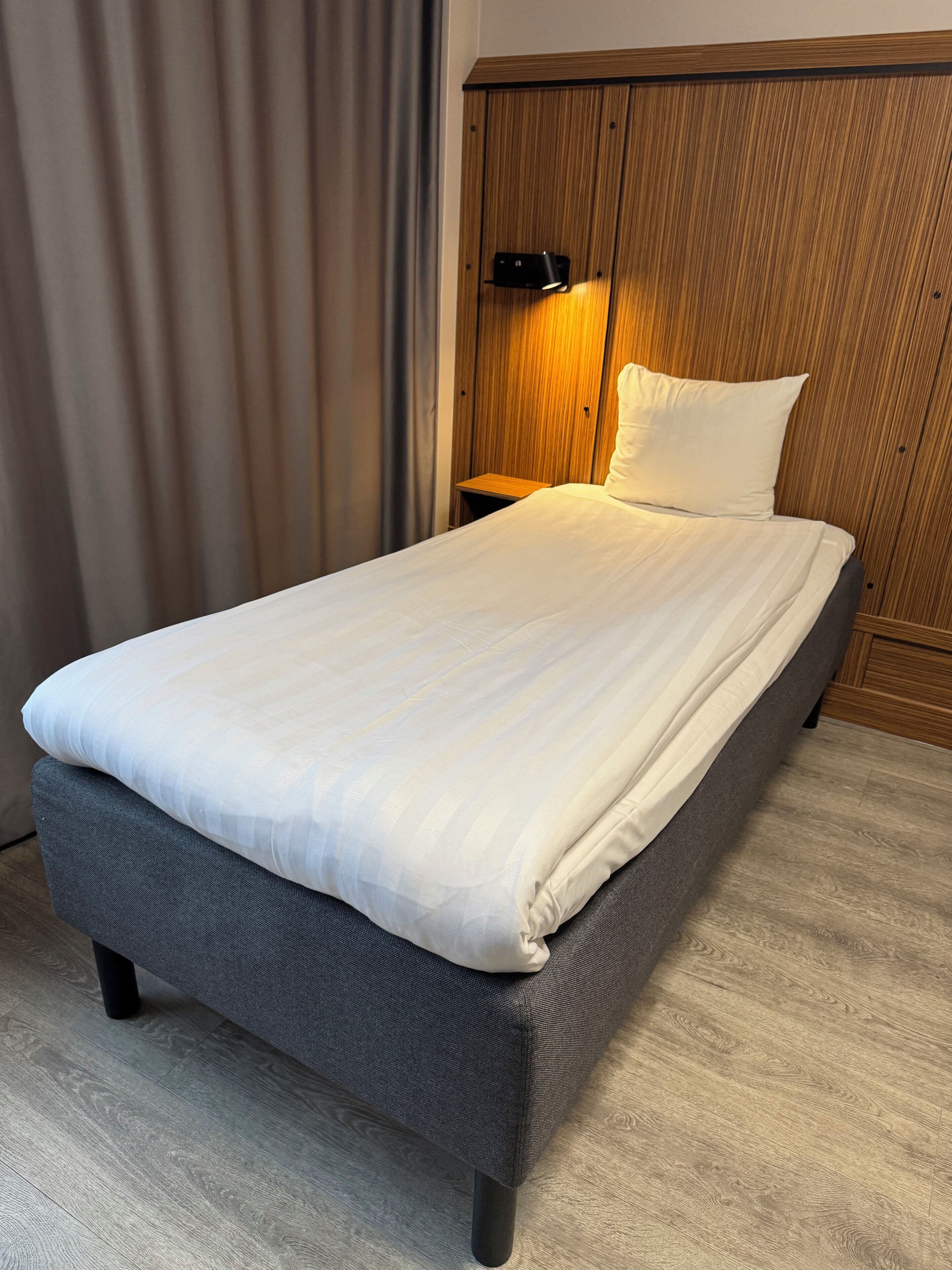 A neatly made bed with a grey headboard, a wooden wardrobe with a mirror, and a small bedside table with a lamp.