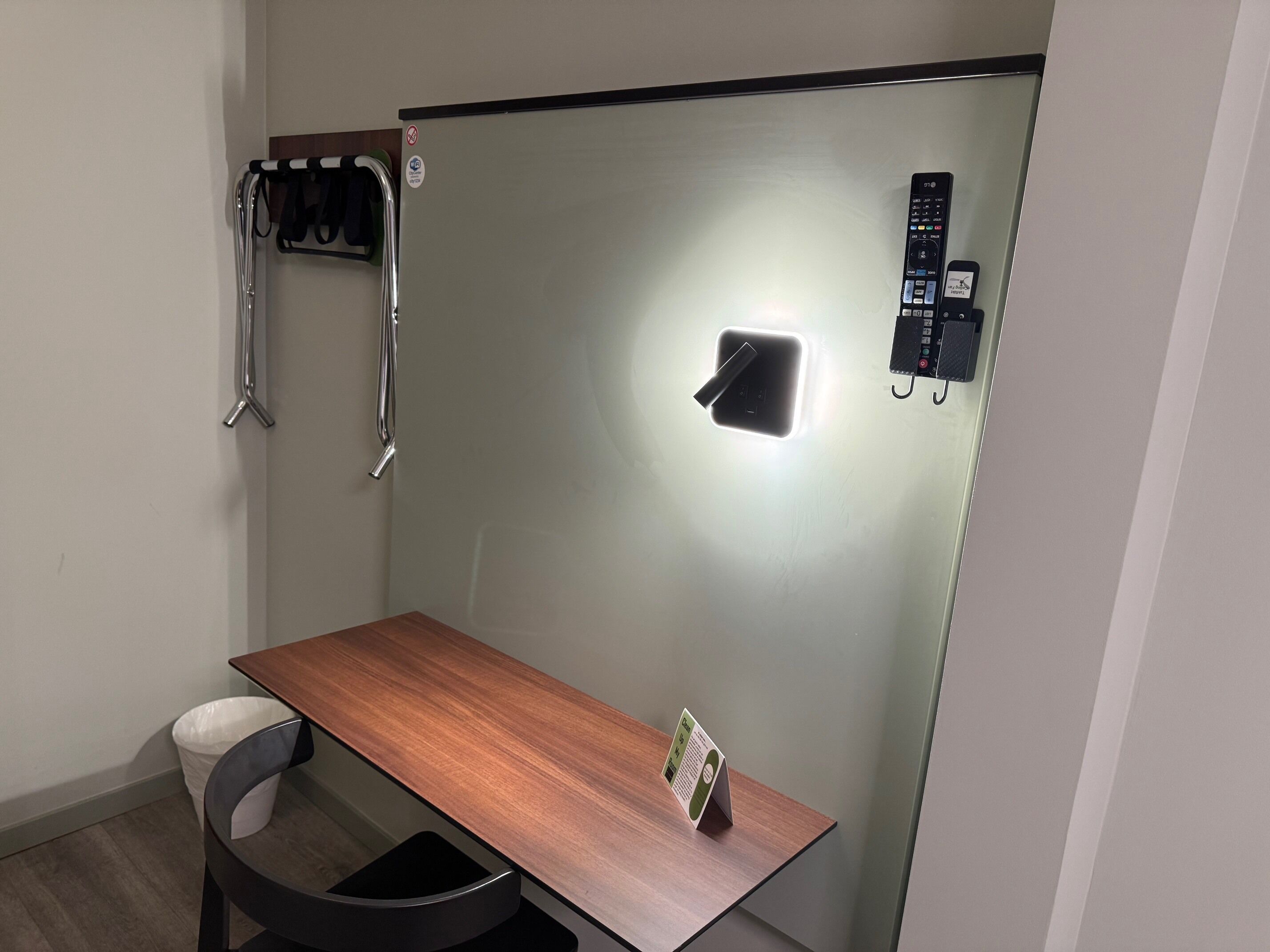 Una habitación pequeña y moderna con un escritorio de madera, una silla, un cesto de basura, un perchero, una lámpara de pared y un teléfono.