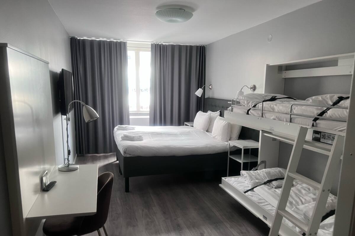 A hotel room with a bunk bed, a desk with a chair, a lamp, and a window with curtains.