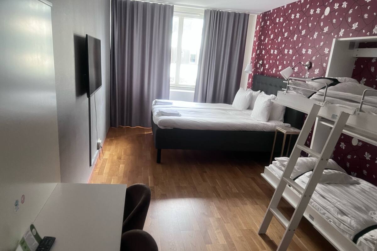 A hotel room with a bed, a desk with a chair, a TV mounted on the wall, and a window with curtains.