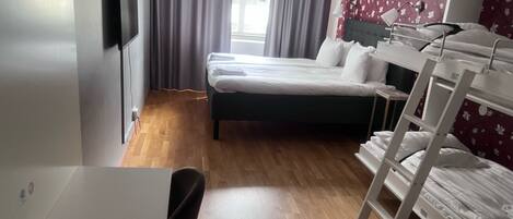 A hotel room with a bed, a desk with a chair, a TV mounted on the wall, and a window with curtains.