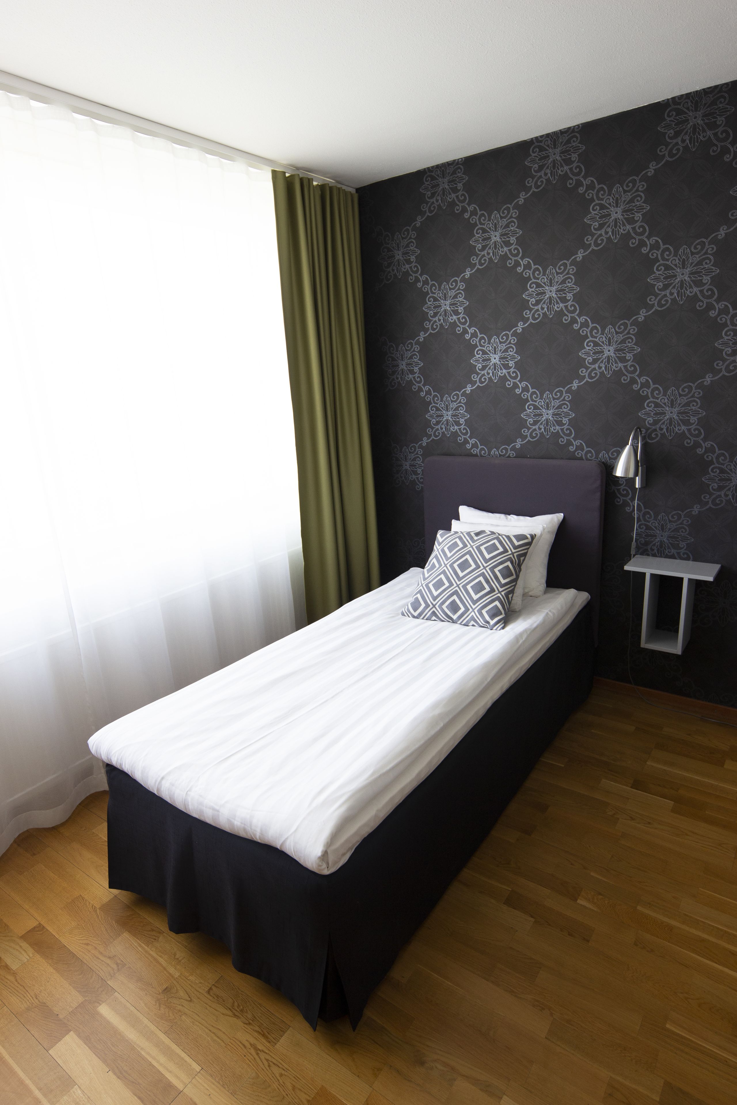 Ein ordentlich bezogenes Bett mit weiß-schwarzer Tagesdecke, einem grünen Vorhang und einer Wand mit Muster.