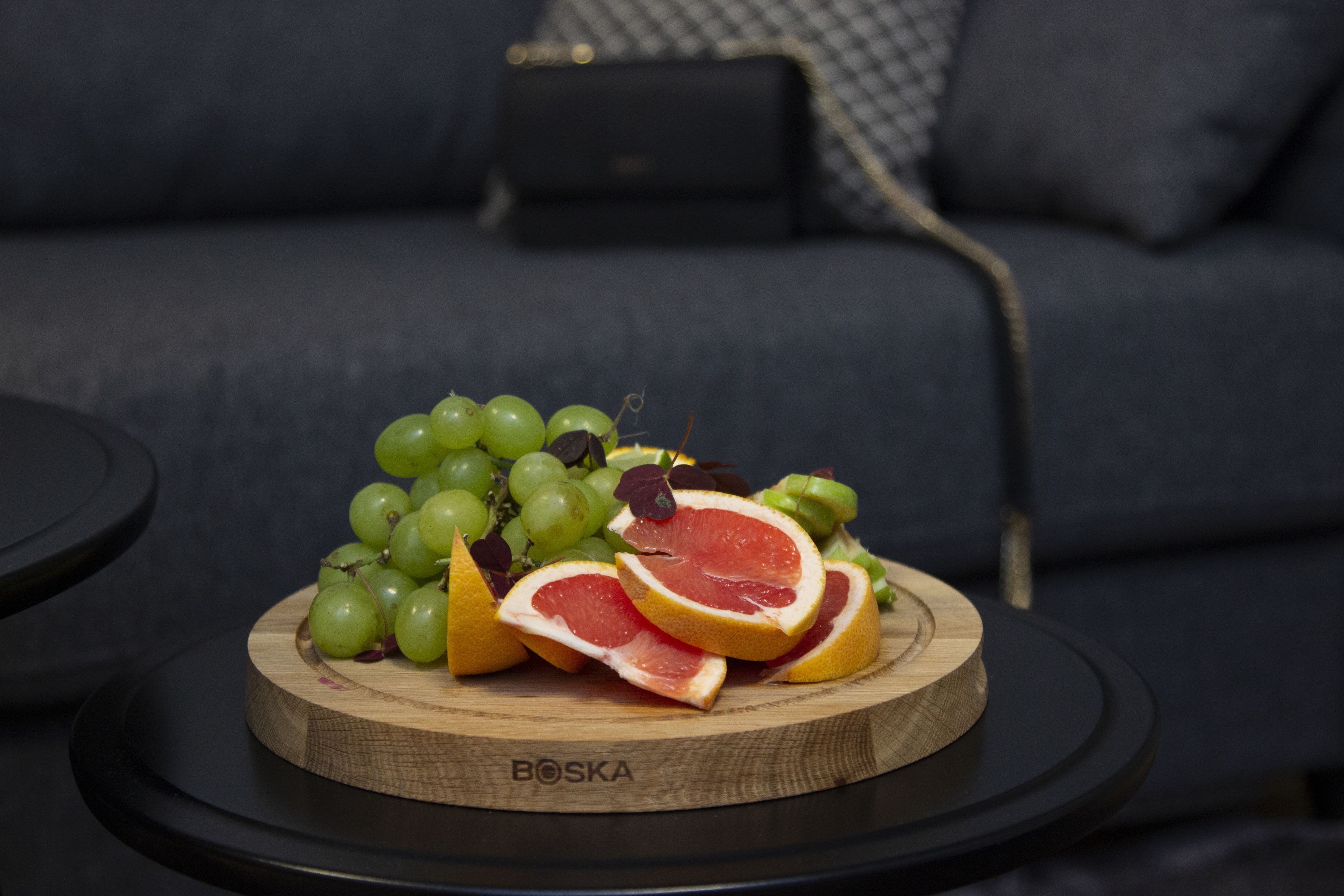 A wooden board with green grapes and halved grapefruit on a round black table, with a dark sofa in the background.