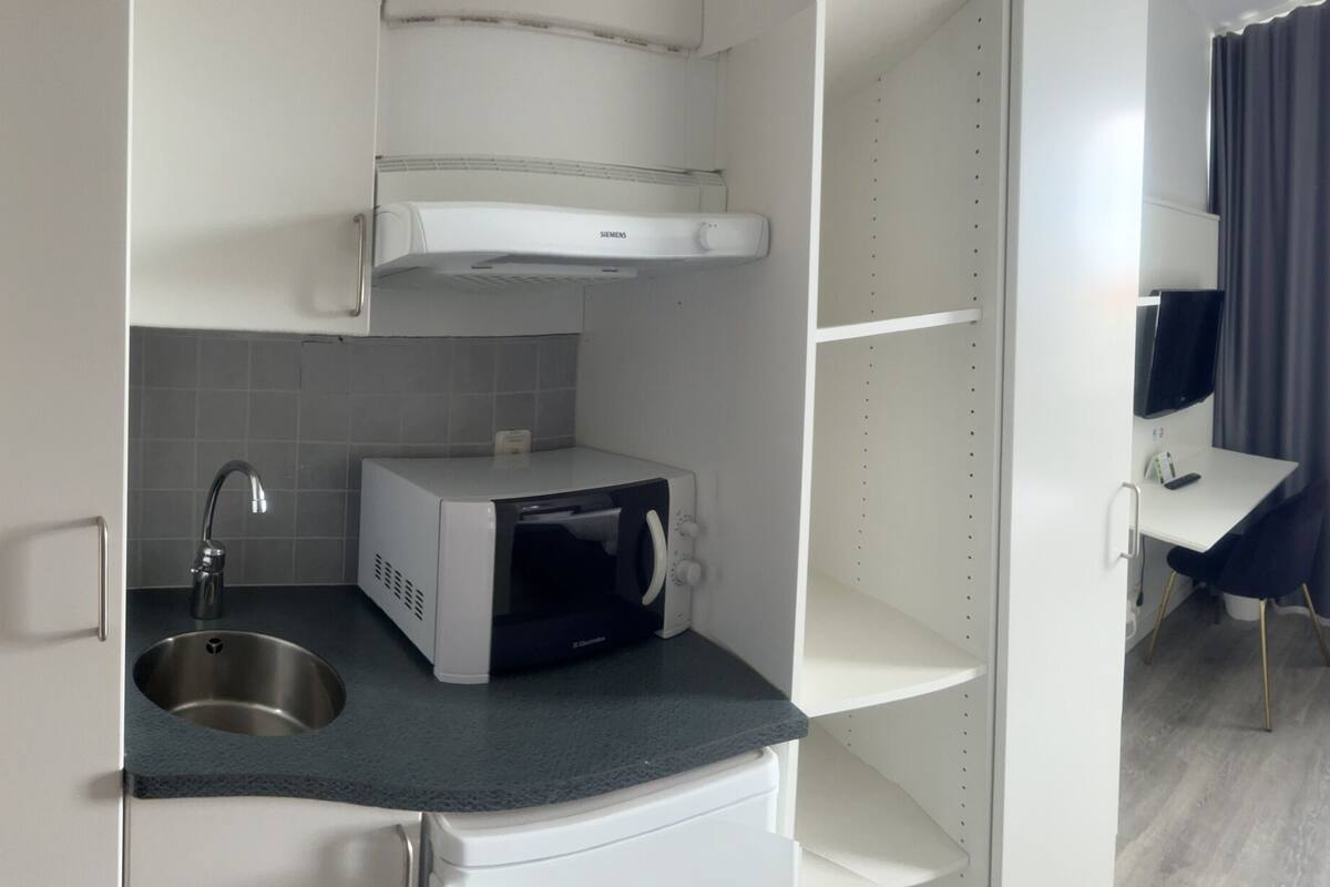A hotel room with a kitchenette, a bed, a desk, and a window with curtains.