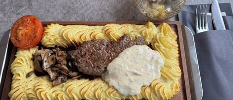 A plated meal with steak, mashed potatoes, and a tomato slice on a wooden board.