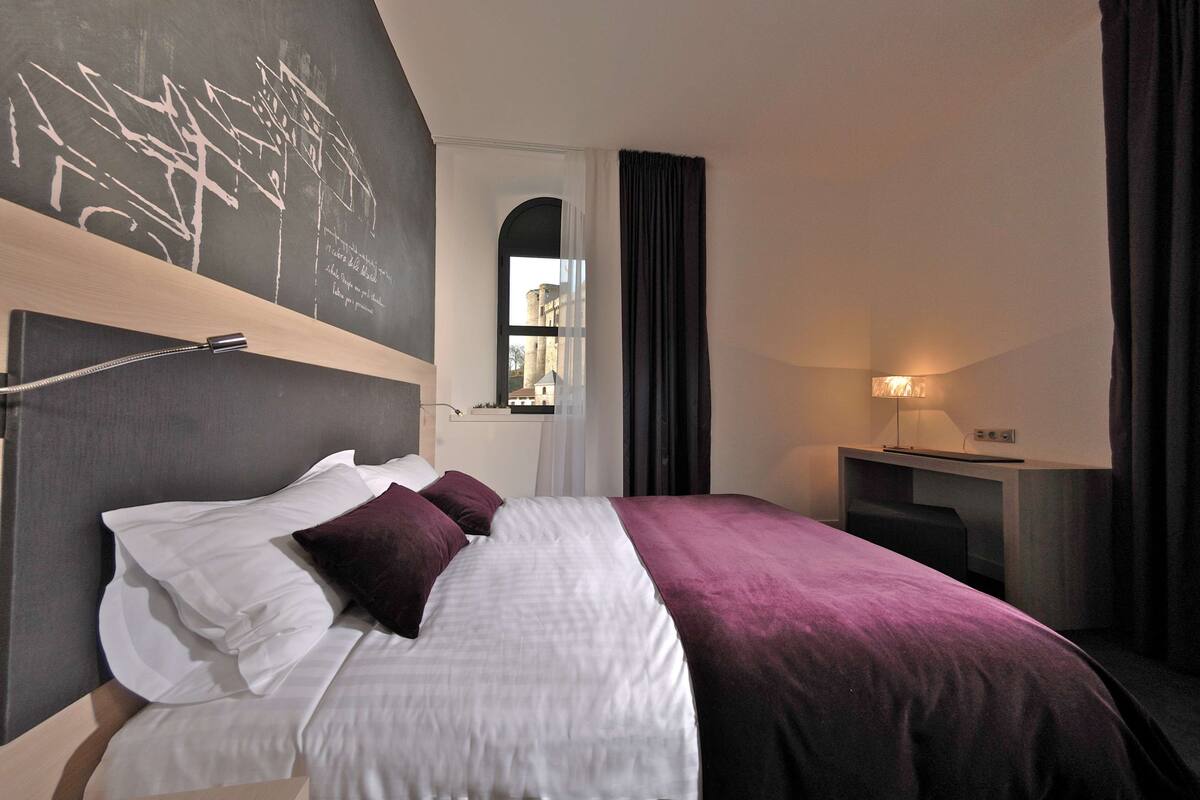 Habitación de hotel con cama, escritorio, lámpara y vista a un edificio a través de una ventana.