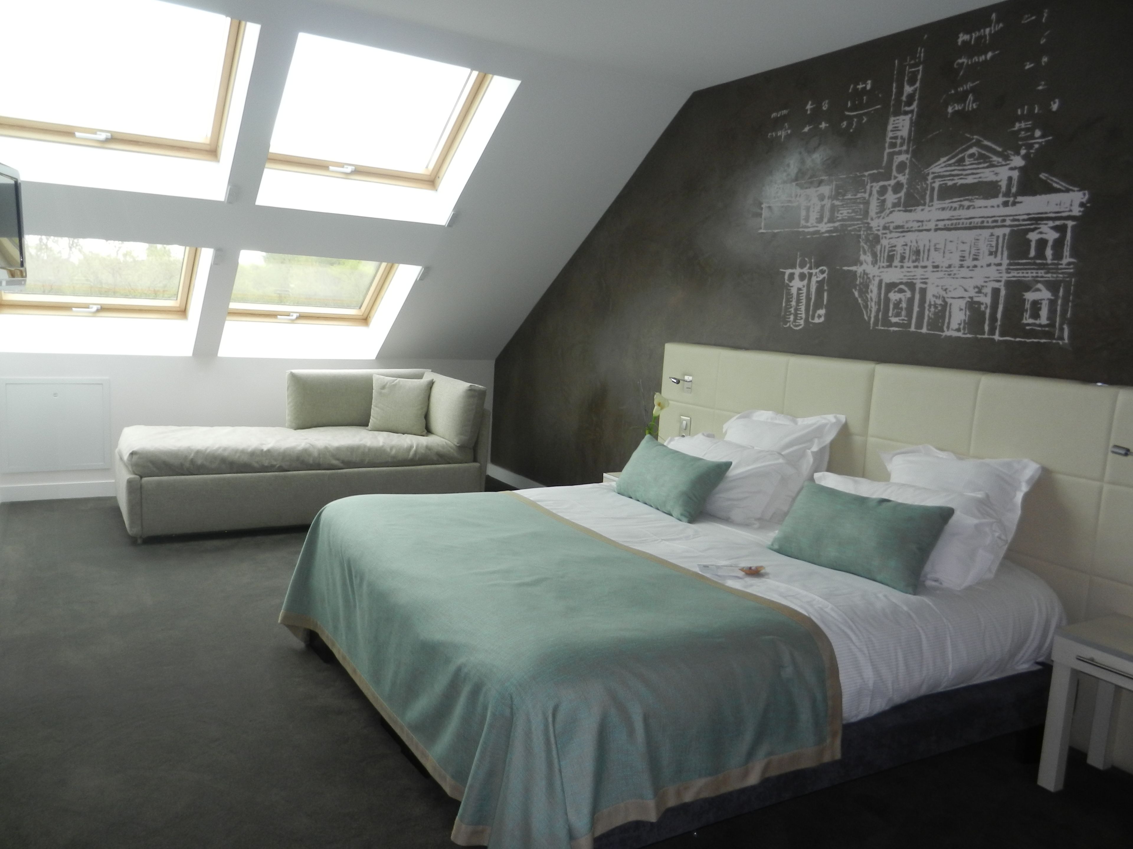 Ein Schlafzimmer mit schrägverdeckter Decke, einem Bett mit grüner Bettwäsche, einer Couch und einer Wandtafel mit architektonischen Zeichnungen.
