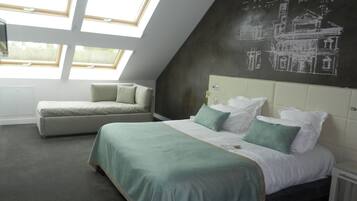 Ein Schlafzimmer mit schrägverdeckter Decke, einem Bett mit grüner Bettwäsche, einer Couch und einer Wandtafel mit architektonischen Zeichnungen.