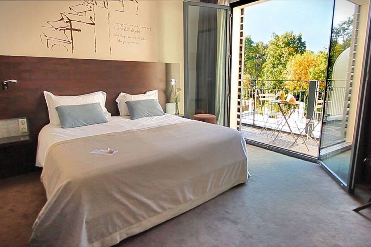 Una habitación de hotel moderna con una cama grande, una mesita de noche y un ventanal que da a un balcón con mobiliario exterior.