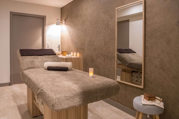 A spa room with a massage table, a small stool, a mirror, and a wall-mounted shelf with towels.