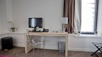 A hotel room with a wooden desk, a clear chair, a television, a lamp, a small plant, and a window with curtains.
