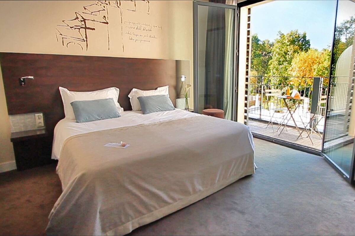 Una habitación de hotel con una cama grande, cabecera de madera, puerta de vidrio que da a un balcón exterior y una pared con detalles decorativos escritos.