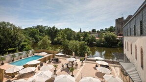Une piscine sur le toit, agrémentée de parasols, de sièges et offrant une vue sur une rivière et une structure ressemblant à un château en arrière-plan.
