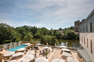 Uma área de piscina no terraço com guarda-sóis, assentos e vista para um rio e uma estrutura semelhante a um castelo ao fundo.