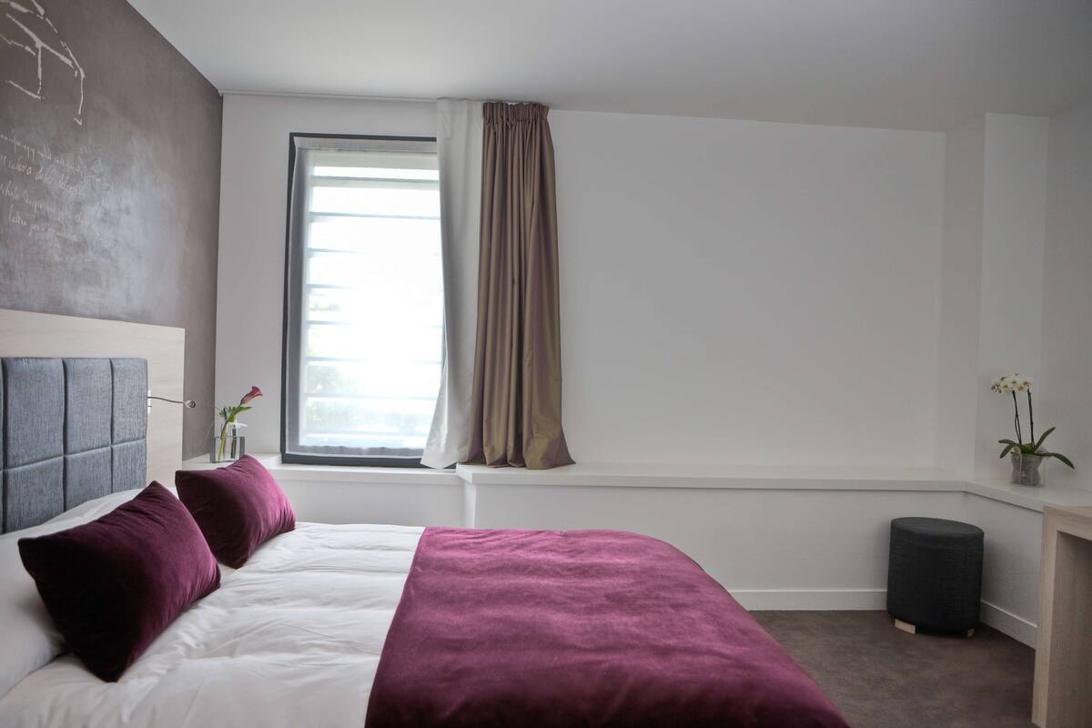 Un dormitorio moderno con una cama grande, una ventana con cortinas, un banquillo redondo pequeño y un jarrón con flores.