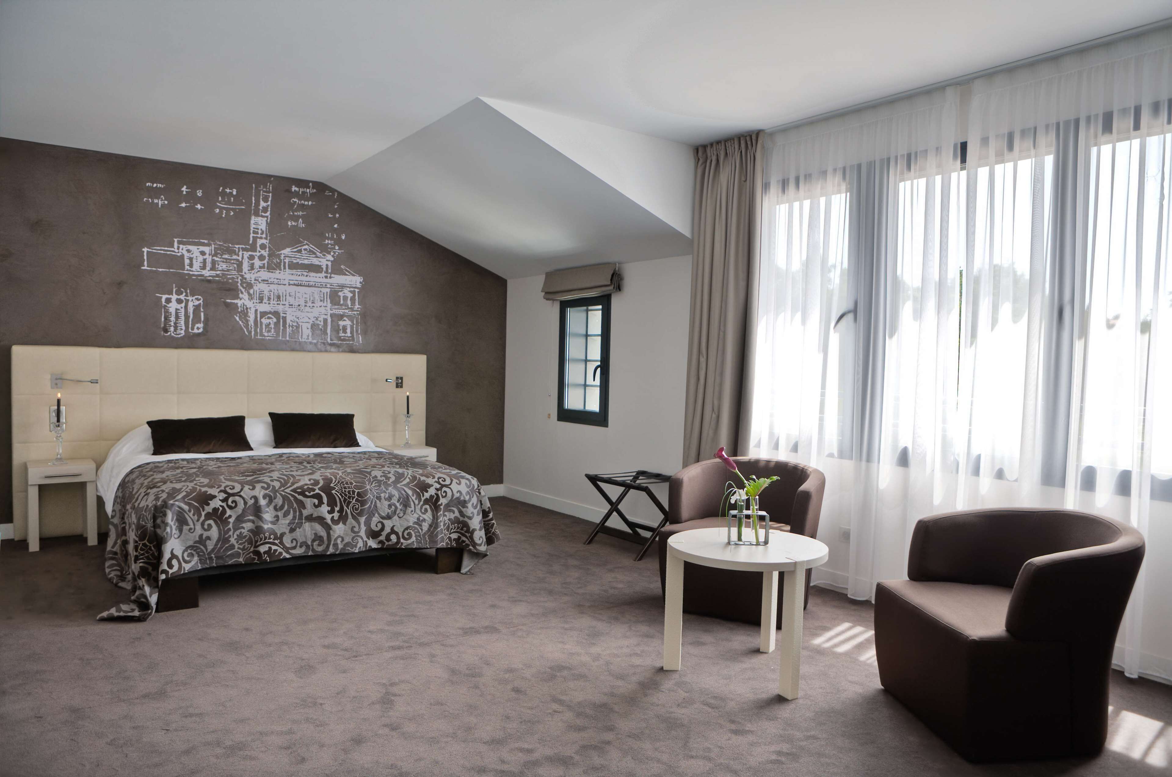 Habitación de hotel con una cama grande, una mesita con un florero, un sillón marrón y una ventana con cortinas translúcidas.