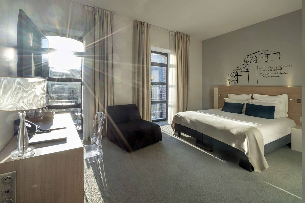 Habitación de hotel con cama, escritorio con lámpara, silla y un amplio ventanal con cortinas.