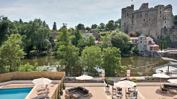 Un espace piscine avec des chaises longues et des parasols, un château en arrière-plan et une rivière.