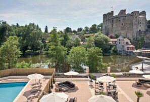 Uma área de piscina com espreguiçadeiras e guarda-sóis, um castelo ao fundo e um rio.