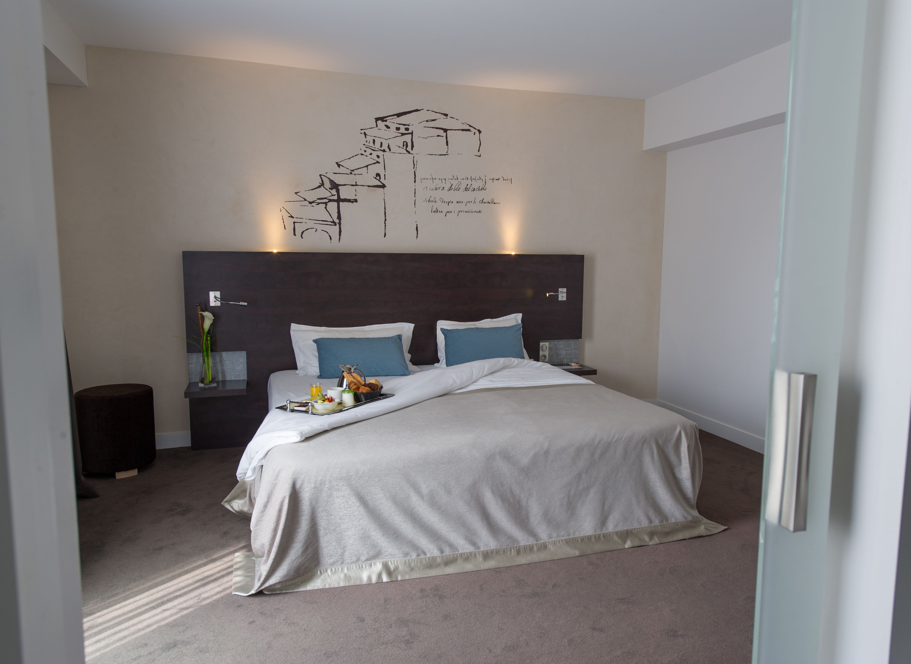 Une chambre d’hôtel moderne avec un grand lit, une table de chevet avec une lampe, une œuvre d’art murale et une petite table d’appoint avec un vase et un plateau contenant des éléments de petit-déjeuner.