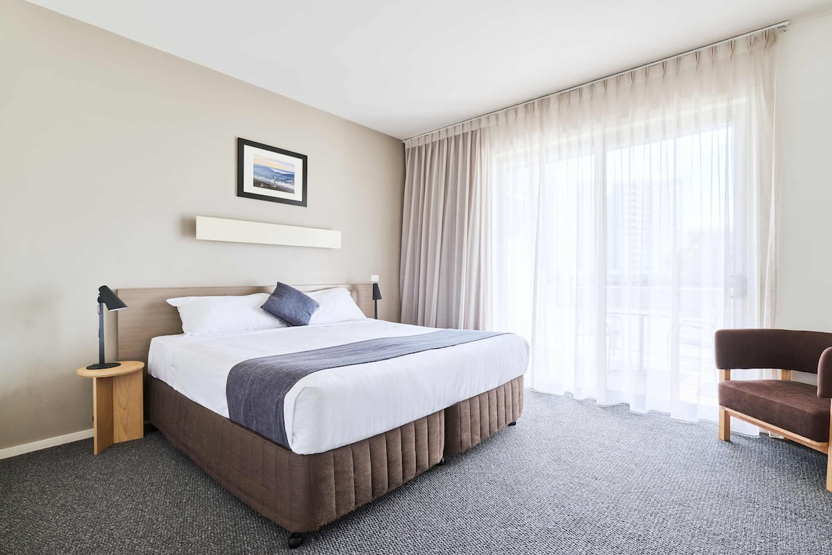 Habitación de hotel con una cama grande, una mesita de noche, una silla, un cuadro en la pared y cortinas de encaje.