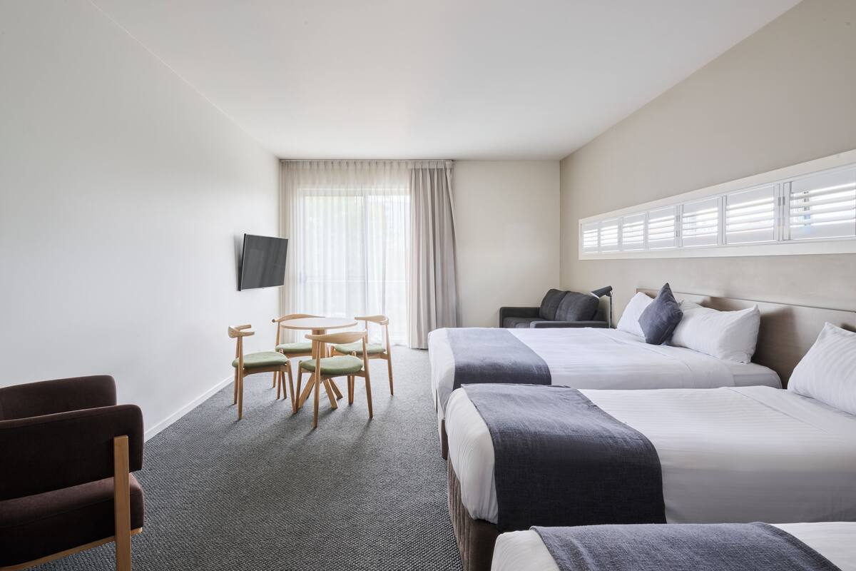 Habitación de hotel con dos camas, un televisor montado en la pared, un escritorio con sillas y una ventana con persianas.