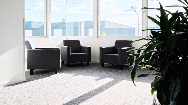 A room with three dark armchairs arranged in a semi-circle, large windows offering a view of buildings, and a potted plant in the foreground. - Abode Gungahlin (Gungahlin)