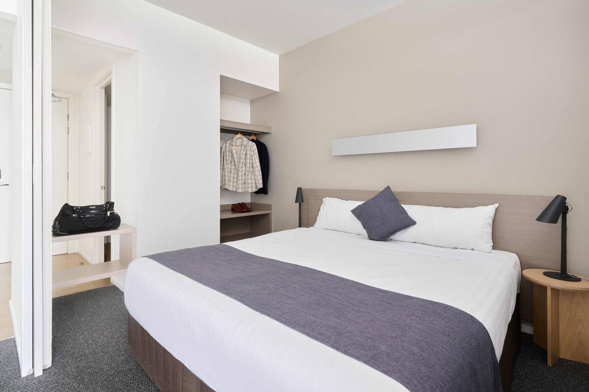 Habitación de hotel ordenada con una cama grande, una mesita de noche con lámpara, un armario y un perchero.
