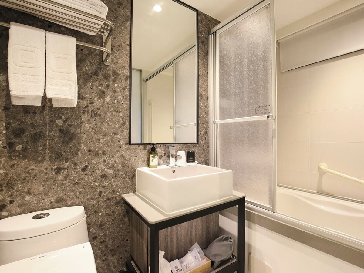 Un baño con pared de mármol, lavamanos blanco, espejo y zona de ducha.