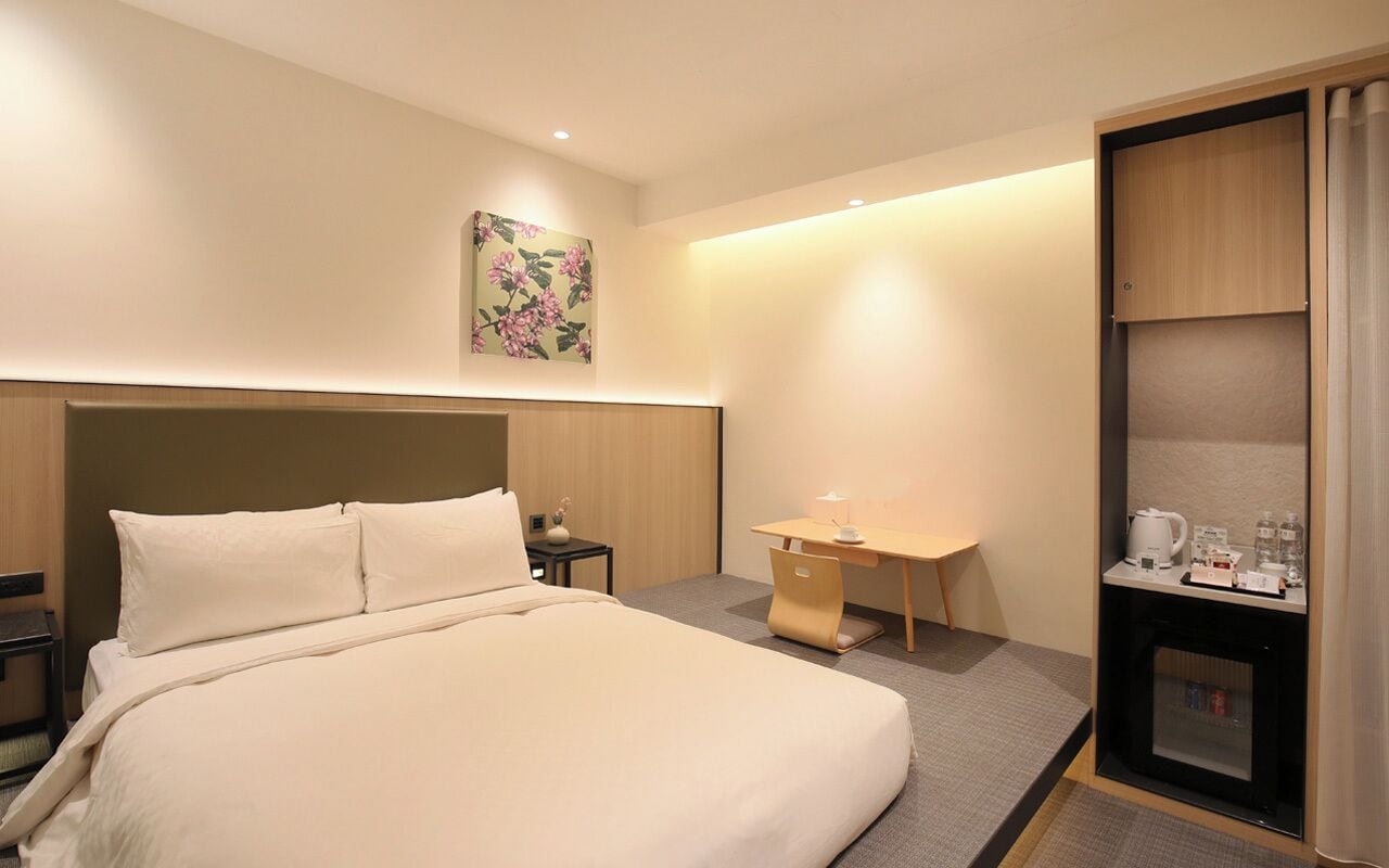 A hotel room with a large bed, a desk, a chair, a small table with a lamp, a picture on the wall, and a minibar.