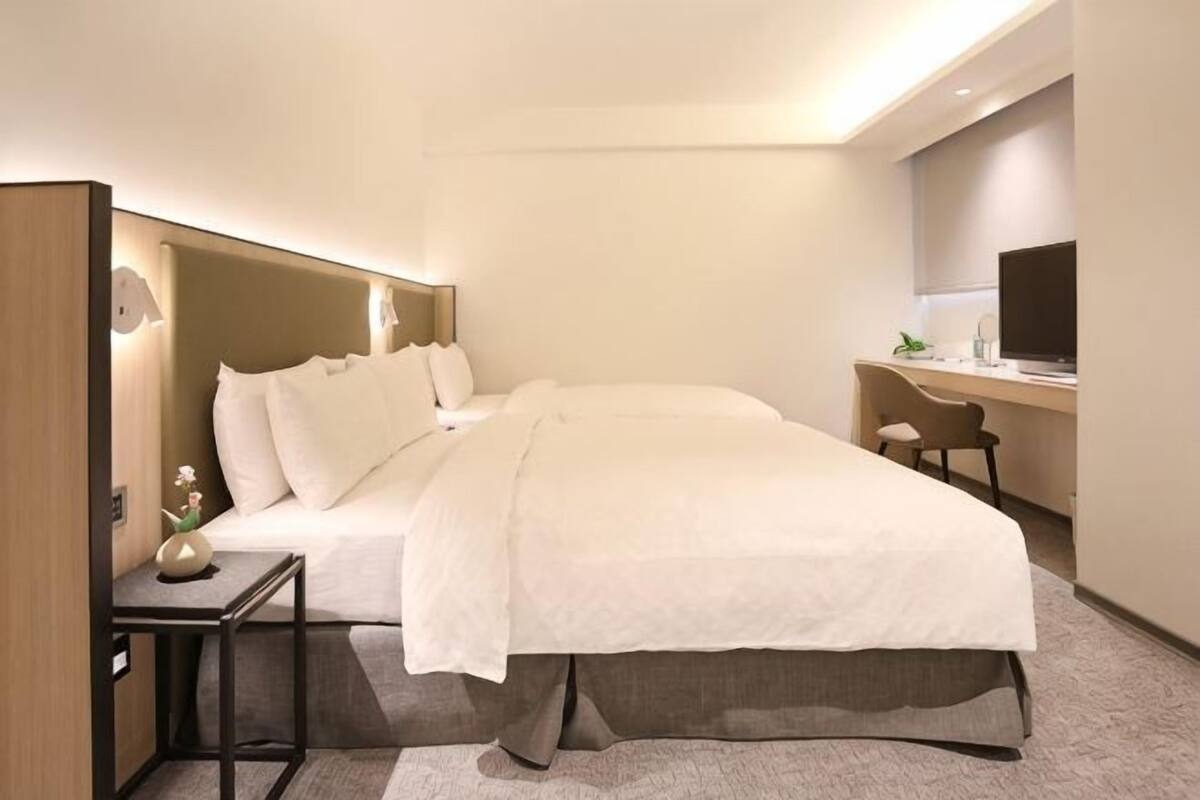 Quarto de hotel com cama, mesa de cabeceira, luminária, escrivaninha, cadeira e televisão.
