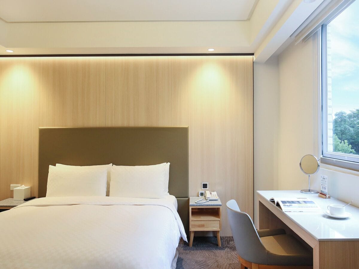 A hotel room with a large bed, a desk with a chair, a bedside table with a phone and a mirror, and a window with a view of trees.