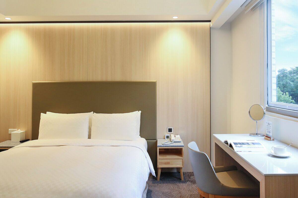 Quarto de hotel com uma cama grande, uma escrivaninha com cadeira, mesa de cabeceira com telefone e espelho, e uma janela com vista para árvores.