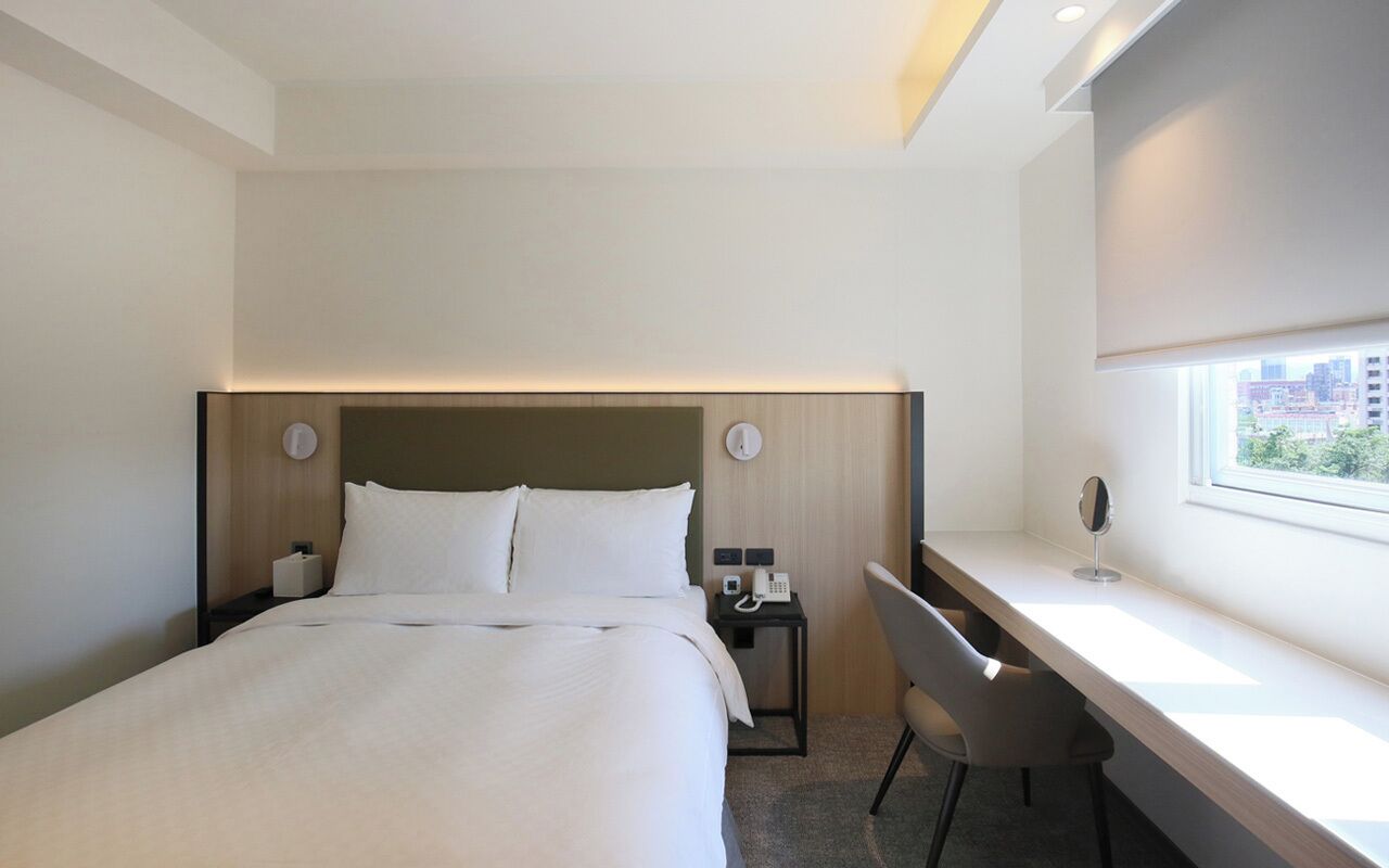 A hotel room with a bed, bedside table with a phone, a desk with a chair, and a window with a view of buildings.