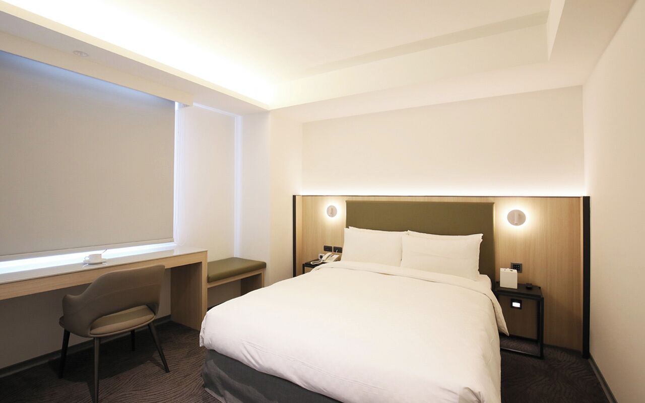 A hotel room with a bed, a desk, a chair, and a nightstand with a phone.