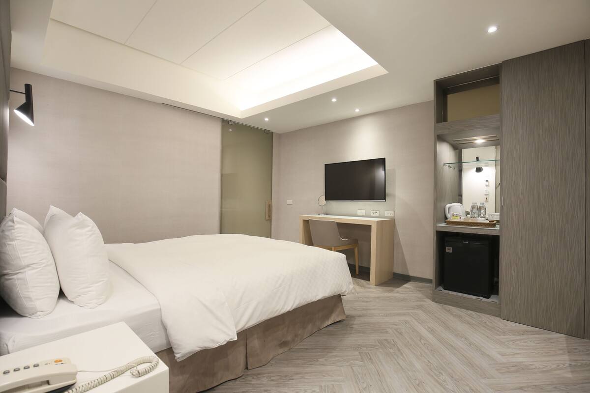 Quarto de hotel moderno com uma cama grande, uma escrivaninha com computador, televisão e uma pequena área de cozinha.