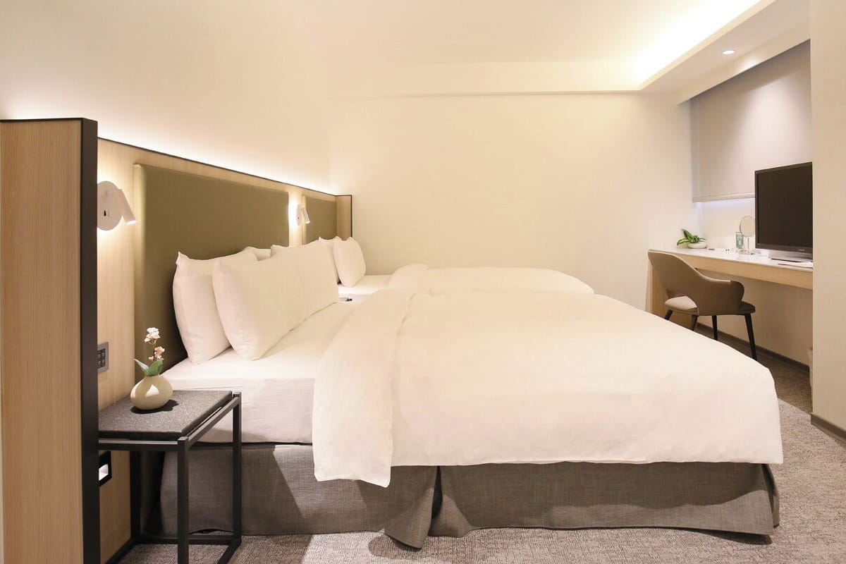 Quarto de hotel com cama, mesa de cabeceira, televisão e escrivaninha.